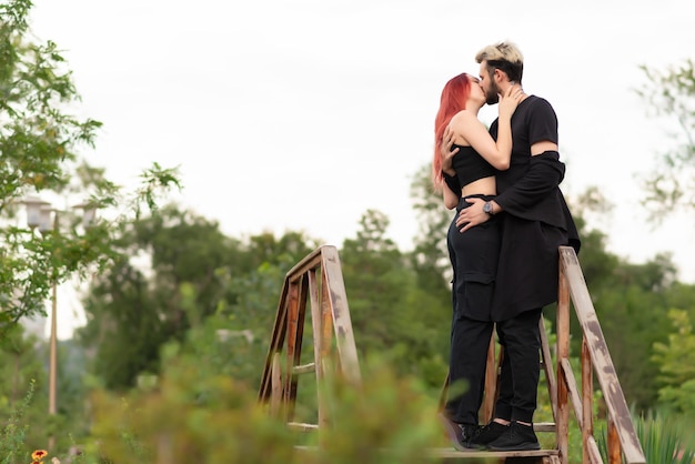 Una pareja elegante enamorada vestida de negro camina por el parque y se abraza Una encantadora pareja de hipsters se besan en una pasarela de madera Disfrutando el tiempo juntos Concepto de amor y estilo de vida juvenil