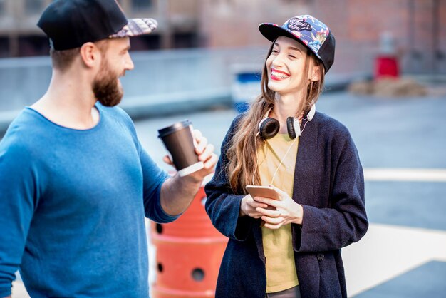 Pareja elegante en suéteres azules y gorras hablando juntos de pie con patineta al aire libre en el patio de recreo de la azotea