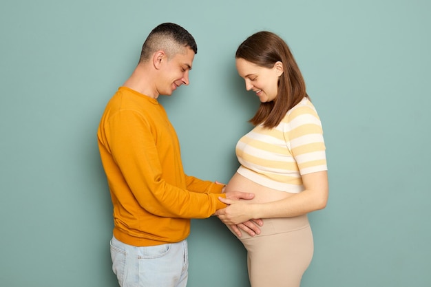 Una pareja embarazada amorosa con ropa casual aislada sobre un fondo azul acariciando el vientre con el futuro bebé tocando el estómago con ternura padres felices que esperan