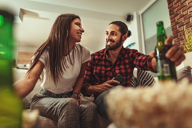 Una pareja emocionada está mirando una película en la televisión en casa sentada en un sofá con palomitas de maíz y cerveza. Tienen un gran fin de semana en buena compañía en el interior.