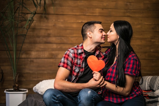 Pareja enamorada besándose con una tarjeta de corazón de papel rojo en casa Beso sensual de una pareja encantadora acostada en la cama mientras celebra el día de San Valentín