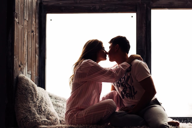 Pareja enamorada en pijama sentada en el alféizar de la ventana y abrazándose