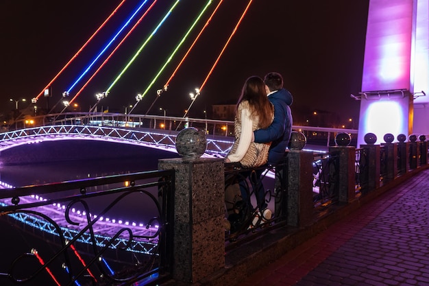 una pareja enamorada se sienta en la barandilla y mira el río cerca del puente con luz de fondo