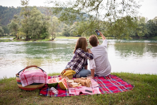 Una pareja enamorada tomándose un selfie por teléfono móvil mientras disfruta de un picnic mientras bebe y come en la hermosa naturaleza en la orilla del río