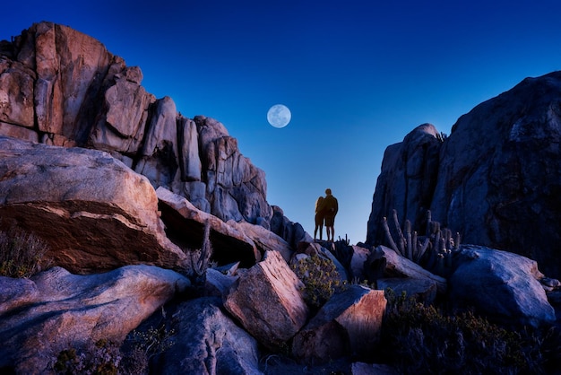 Pareja de enamorados enormes en la roca mirando el cielo nocturno con la luna sobre las montañas