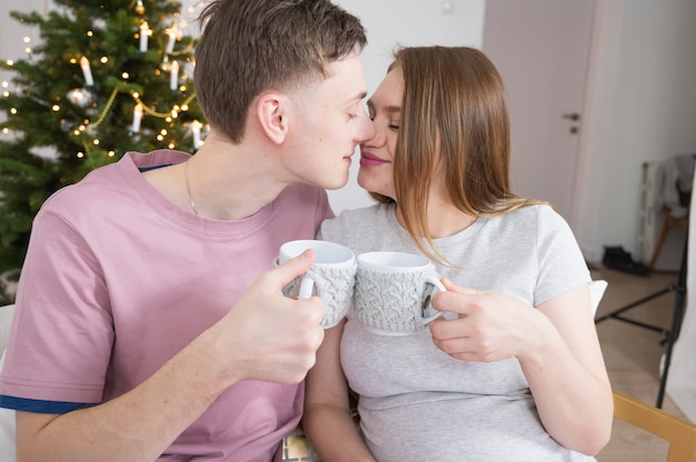 Pareja de enamorados hombre y mujer bebiendo té café sentado en un sofá con decoración navideña en segundo plano.