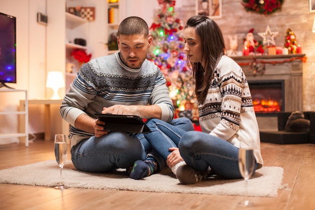 Pareja de enamorados sentados delante del árbol de Navidad mirando en la computadora de la tableta.