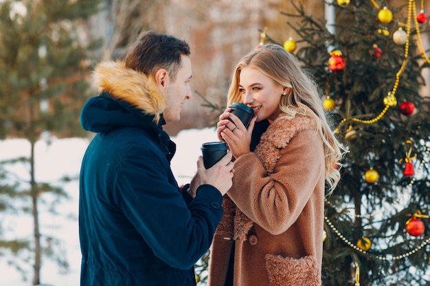 Pareja de enamorados tomando café junto al árbol de Navidad decorado en el bosque de invierno.