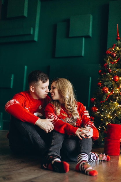 Una pareja encantadora enamorada con suéteres rojos tradicionales de Navidad pasa tiempo cerca del árbol de Navidad