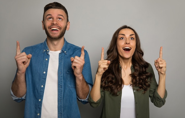 Foto una pareja encantadora posa sobre un fondo gris, vestida con camisetas blancas y camisetas de colores, apuntando hacia la izquierda con las manos y sonriendo a la cámara.