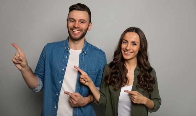 Una pareja encantadora posa sobre un fondo gris, vestida con camisetas blancas y camisetas de colores, apuntando hacia la izquierda con las manos y sonriendo a la cámara.