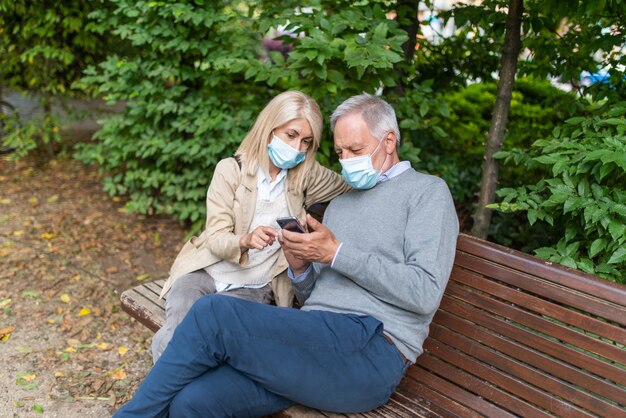 Pareja enmascarada usando un teléfono móvil juntos en el parque durante la pandemia de coronavirus