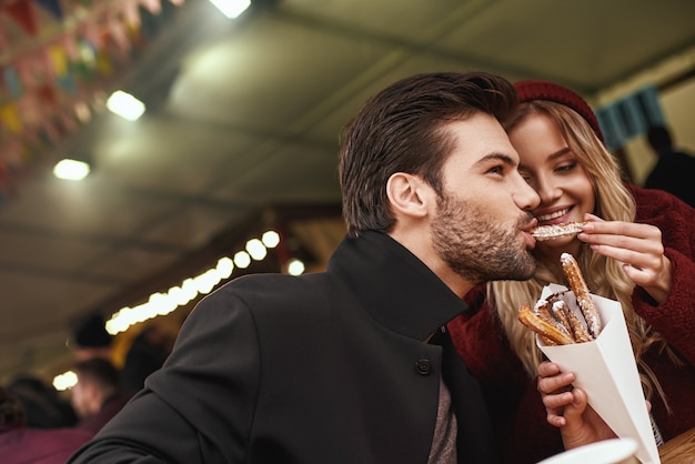 La pareja está comiendo al aire libre. Pareja joven está comiendo churros en el mercado de comida de la calle. Temporada de frio. Foto de primer plano de la pareja comiendo de un paquete