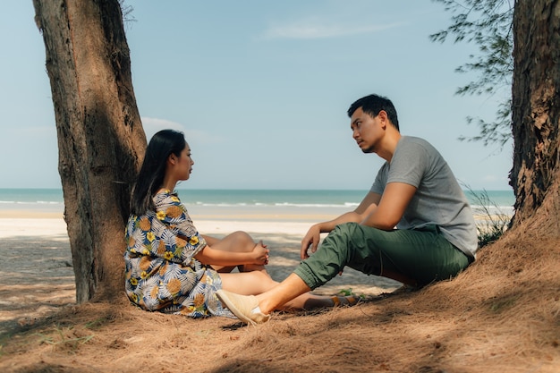 Pareja están sentados juntos en la playa bajo el árbol de pino.