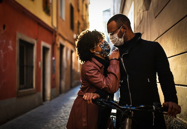 Pareja étnica negra con máscara protectora se besan al atardecer en bicicleta