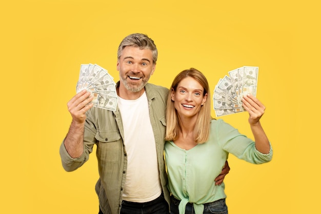 Una pareja europea de mediana edad llena de alegría sosteniendo fanáticos del dólar en efectivo sonriendo a la cámara en un estudio amarillo