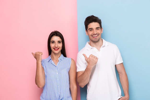 Pareja europea en ropa casual sonriendo y haciendo gestos con los dedos a un lado, aislado sobre pared colorida