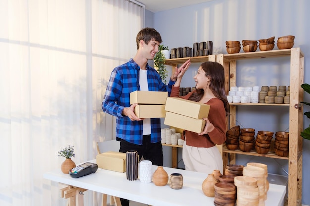 La pareja de éxito de hombre caucásico y mujer asiática venden productos de jarrón en línea en vivo en casa de pie alto cinco sosteniendo paquetes para enviar por orden a los clientes concepto de compras en línea