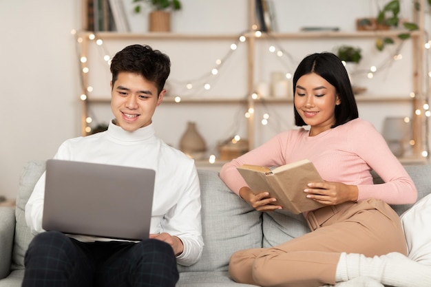 Pareja de familia asiática relajante con portátil y libro sentado en el sofá en casa por la noche. Pareja y relación romántica. Ocio de fin de semana hogareño, comodidad doméstica y concepto de relajación