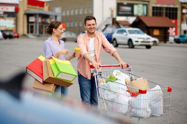Pareja de familia con bolsas en el carro en el estacionamiento del supermercado. Clientes felices con compras en el centro comercial, vehículos en segundo plano.