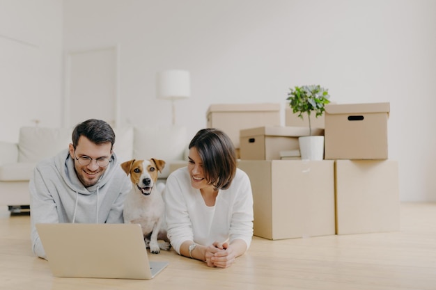 Una pareja familiar toma un descanso en su nuevo apartamento después de mudarse en el piso y navegar por Internet a través de una computadora portátil posada en una espaciosa sala luminosa con lámpara de sofá y un montón de cajas de cartón para mascotas