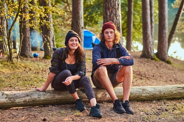 Una pareja feliz y atractiva se relaja en el bosque verde y soleado mientras se sienta en el tronco. Hay camping en el fondo.