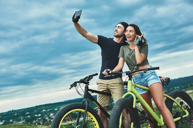 Pareja feliz con bicicleta tomando selfie por teléfono inteligente al aire libre