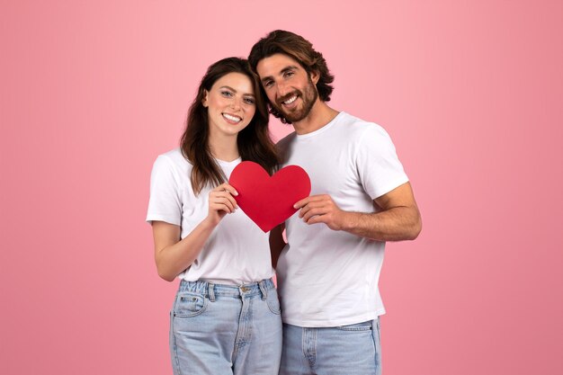 Una pareja feliz con camisetas blancas y vaqueros sosteniendo un corazón de papel rojo juntos