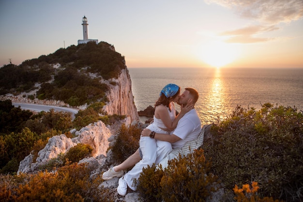 Una pareja feliz conociendo la puesta de sol sentada en el acantilado con el faro de Lefkada al fondo