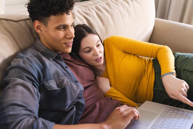 Una pareja feliz y diversa acostada en el sofá y usando una computadora portátil en la sala de estar en el espacio de copia de casa
