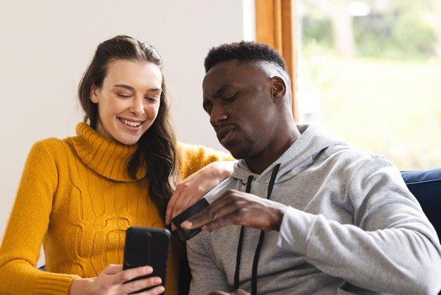 Una pareja feliz y diversa sentada en el sofá usando un teléfono inteligente para comprar en línea en casa