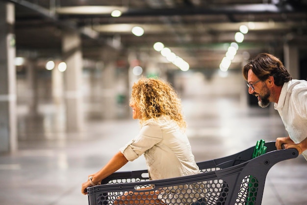 Pareja feliz divirtiéndose con carrito de compras en el estacionamiento Esposo empujando a su esposa despreocupada dentro del carrito de compras La pareja se divierte con carrito de supermercado Concepto de economía familiar