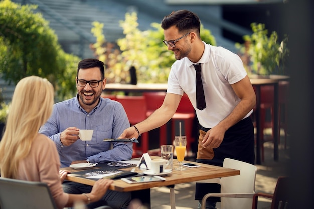 Una pareja feliz hablando mientras el camarero prepara la mesa para cenar y les da cubiertos en un restaurante