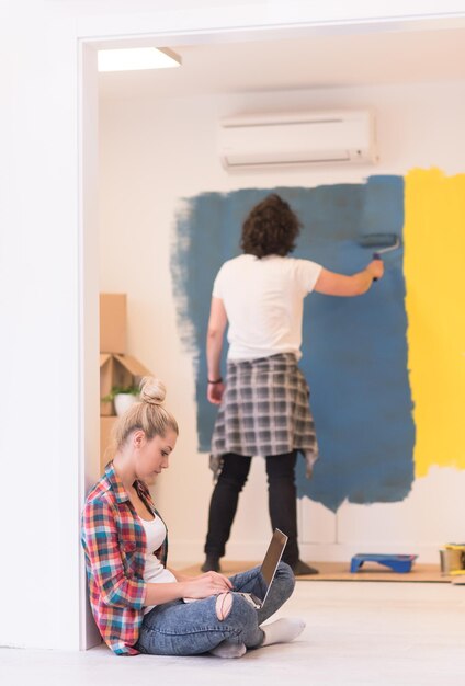 Pareja feliz haciendo renovaciones en el hogar, el hombre está pintando la habitación y la mujer se relaja en el suelo y se conecta con una computadora portátil