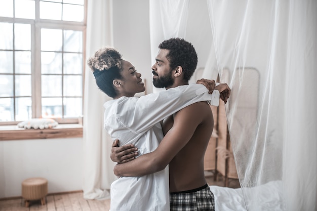 Pareja feliz. Hombre barbudo joven de piel oscura y bonita esposa abrazándose mirando el uno al otro en casa en la mañana