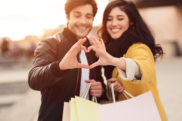 Pareja feliz mostrando el signo del corazón con las manos sosteniendo bolsas de compras