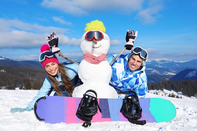 Pareja feliz con muñeco de nieve y snowboard en la estación de esquí. Vacaciones de invierno