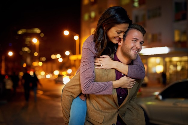 Pareja feliz y piggyback en la ciudad con el amor por la noche para el vínculo romántico y la fecha del aniversario al aire libre la gente sonríe y una relación saludable en la calle con el paseo nocturno cuidado y apoyo en la ciudad