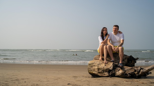 Pareja feliz posando en madera a la deriva cerca del mar Pareja amorosa abrazándose durante la cita en la playa contra el mar agitado y el cielo despejado