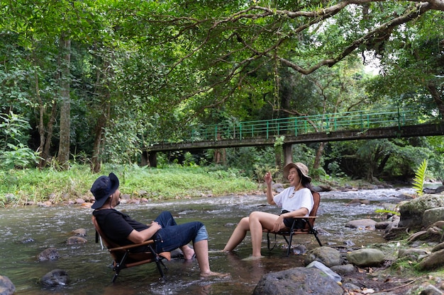 Una pareja feliz sentada en una silla de madera rodeada de vegetación charlando felizmente los fines de semana hablando felizmente en medio del arroyo con los pies en el agua felizmente