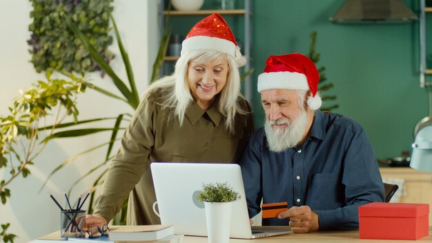 Foto pareja feliz con sombreros de santa claus está eligiendo regalos de navidad compras en línea