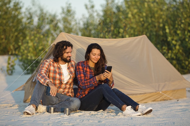 Pareja feliz con un teléfono inteligente en el campamento cerca de la tienda al aire libre