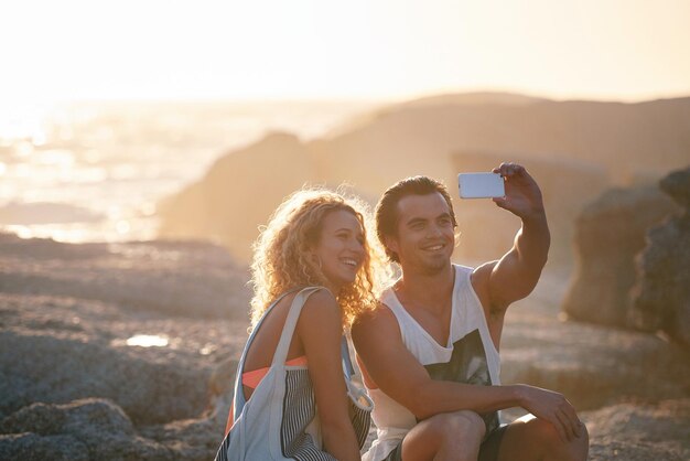 Pareja feliz tomando fotos usando un teléfono inteligente en la playa compartiendo vacaciones románticas juntos