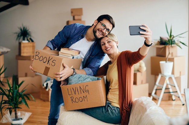 Una pareja feliz tomándose selfie con un teléfono inteligente mientras se muda a un nuevo hogar
