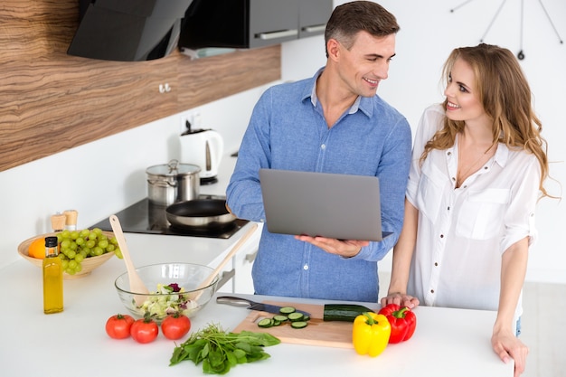 Pareja feliz usando laptop y preparando platos vegetarianos en la cocina de casa