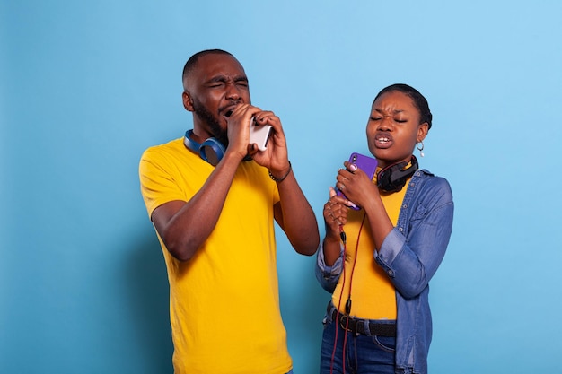 Pareja feliz usando un teléfono inteligente como micrófono para cantar una canción, usando auriculares en el estudio. Hombre y mujer modernos disfrutando de una sesión de karaoke con música mp3, sosteniendo teléfonos móviles en la mano.