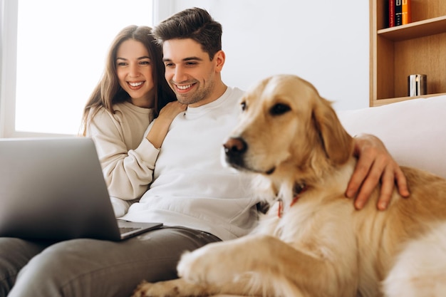 Una pareja feliz viendo una computadora portátil con un perro golden retriever en el sofá en casa