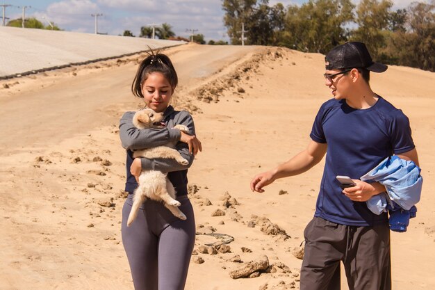 Pareja de fitness caminando sobre la arena mientras la niña lleva a su cachorro en brazos.