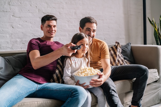 Una pareja gay disfruta viendo películas con su hija sentados juntos en un sofá en casa. Concepto de familia.