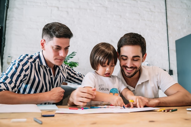 Una pareja gay se divierte con su hijo mientras dibuja algo en un papel en casa. Concepto de familia.